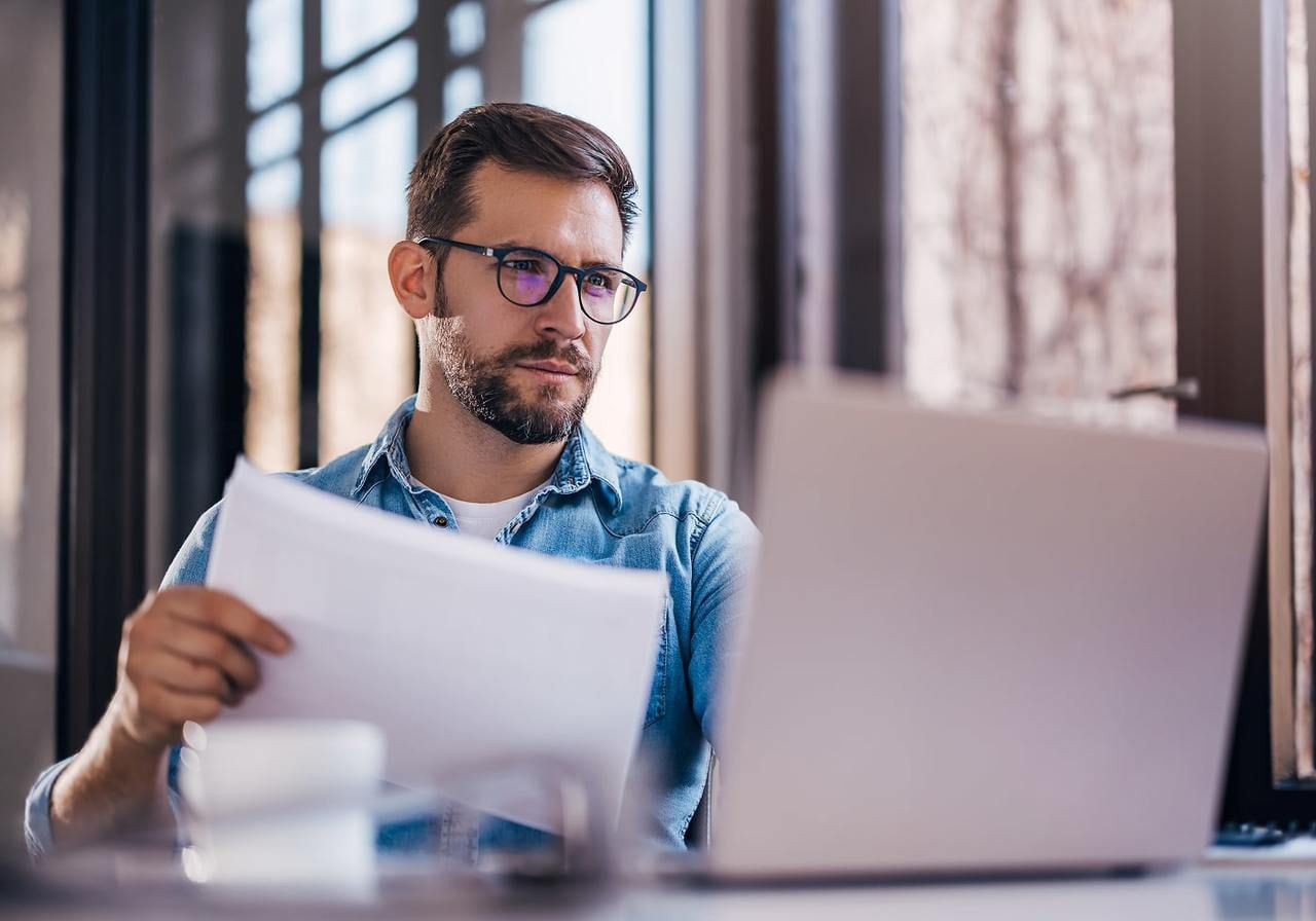 A man in glasses is holding a piece of paper and reading “What is dropshipping?” from a laptop.
